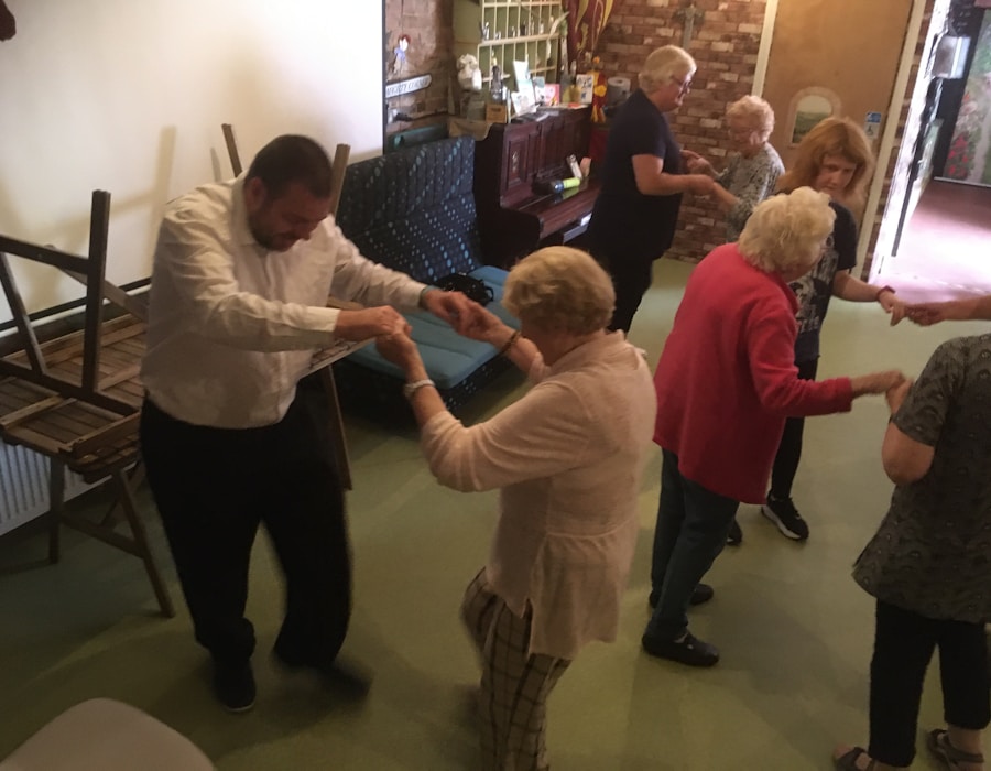 A group of elderly people are engaging in a dance activity in a room. Some chairs are stacked on the side, and a piano is visible in the background. The atmosphere appears lively and social.