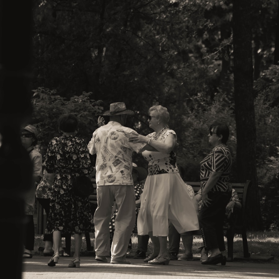 A group of elderly people is gathered in a park, engaging in social interaction. Some are dancing while others watch and converse. The setting is shaded by trees, creating a peaceful and intimate atmosphere.